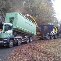 Abrollcontainer mit offenem Aufbau aus der Serie EKO2 in Grün, mit Tunnel und montiertem Hakengerät, wird gerade mit Holzschnitzeln beladen